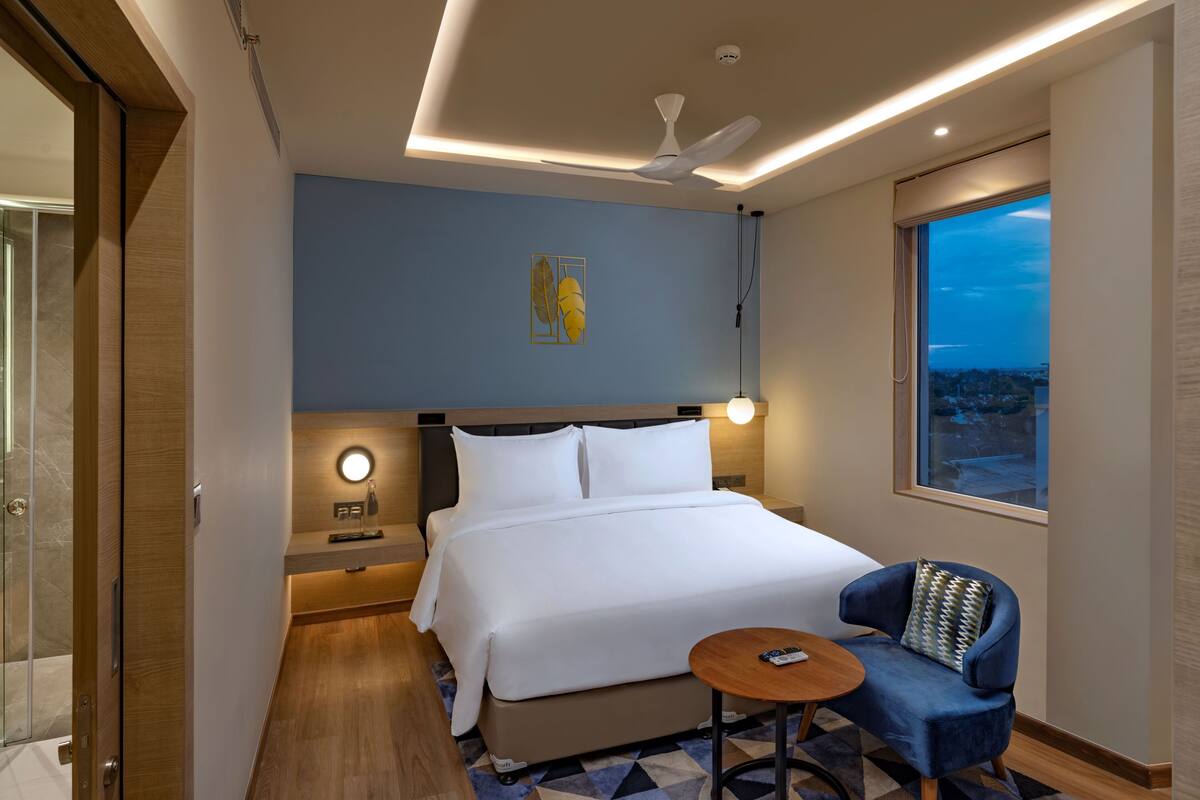 A hotel room with a bed, a chair, a small table, a nightstand, a ceiling fan, a window with a view, and a wall art piece.