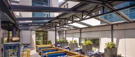 A modern outdoor dining area with wooden benches and tables, glass partitions, and potted plants.