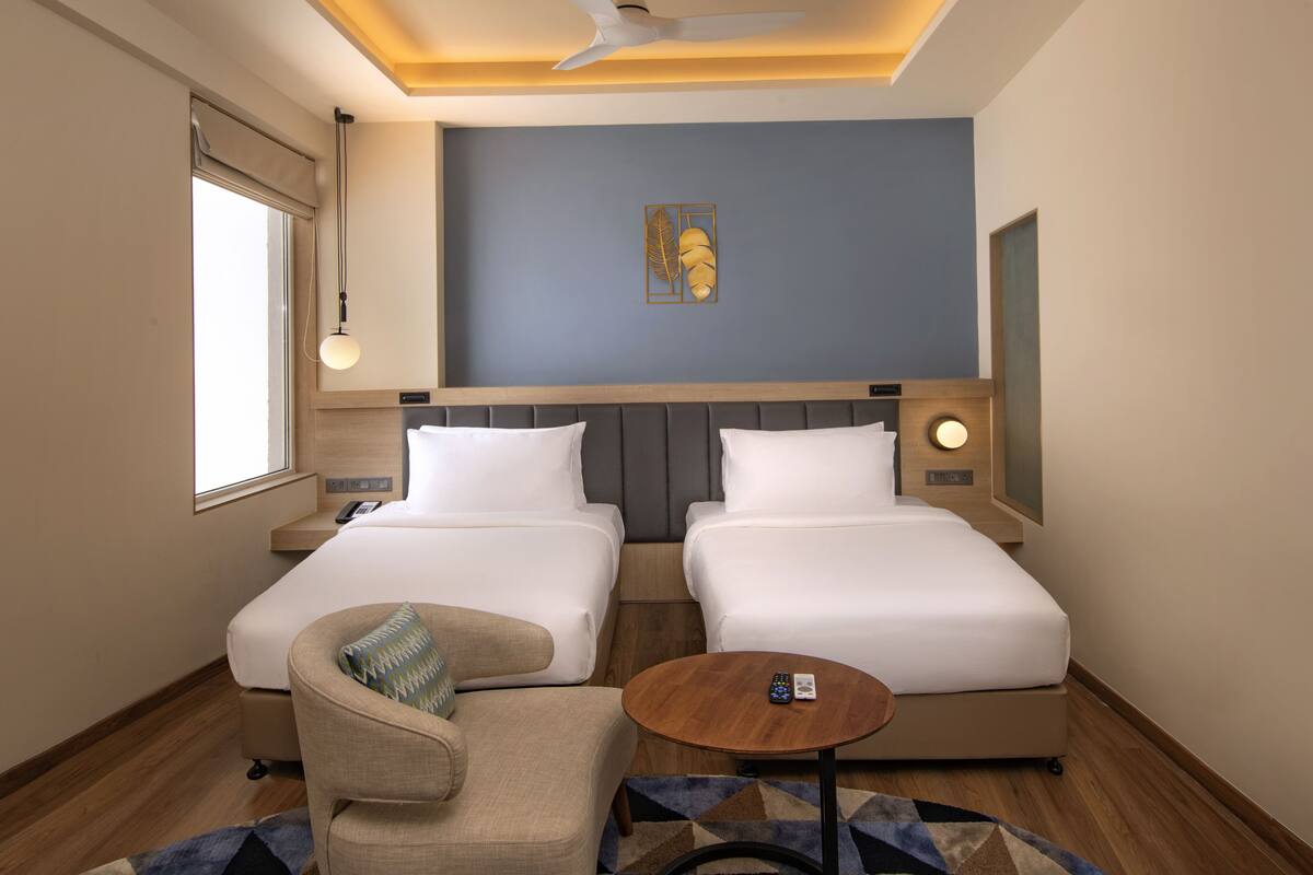 A hotel room with two beds, a chair, a small table, a ceiling fan, a window with a blind, and a wall-mounted artwork.
