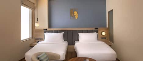 Habitación de hotel con dos camas, una silla, una mesita, un ventilador de techo, una ventana con persiana y una obra de arte colgada en la pared.