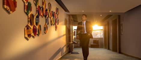 A man walking down a hallway with a wall of colorful ties. The hallway has a carpeted floor and a ceiling with recessed lighting.