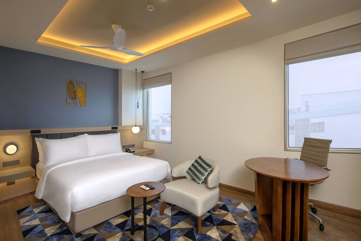 A modern hotel room with a large bed, a small sofa, a round wooden table, and a view of the city through the window.