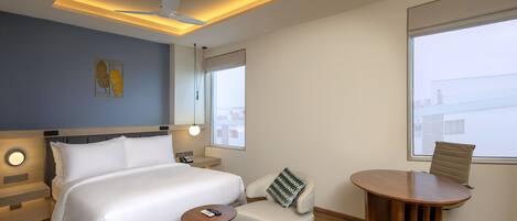 A modern hotel room with a large bed, a small sofa, a round wooden table, and a view of the city through the window.
