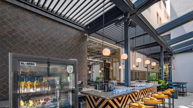 A modern bar area with a glass display case of desserts, a counter with bar stools, and a view of the interior space. - O by Tamara Coimbatore (Coimbatore)