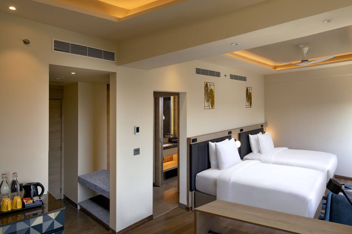 A hotel room with two beds, a desk, a coffee station, and a bathroom visible through an open door.