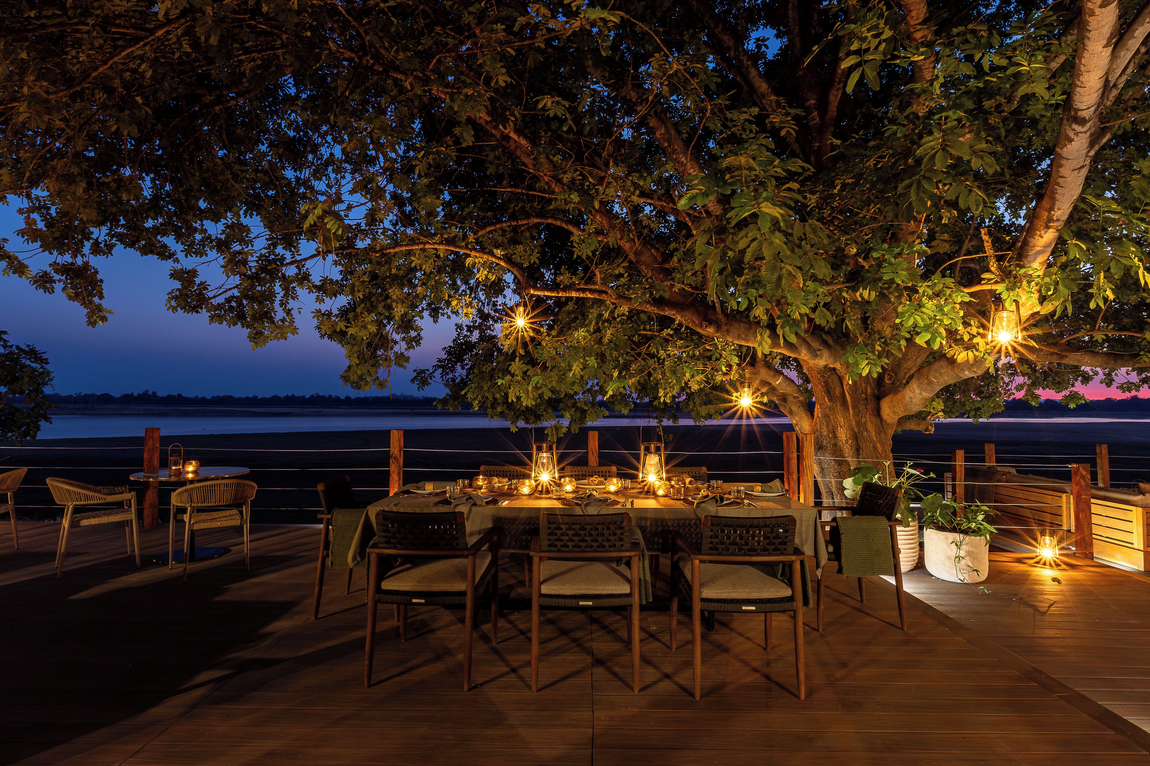 An outdoor dining area with a table set for two, surrounded by chairs, under a tree with hanging lights.