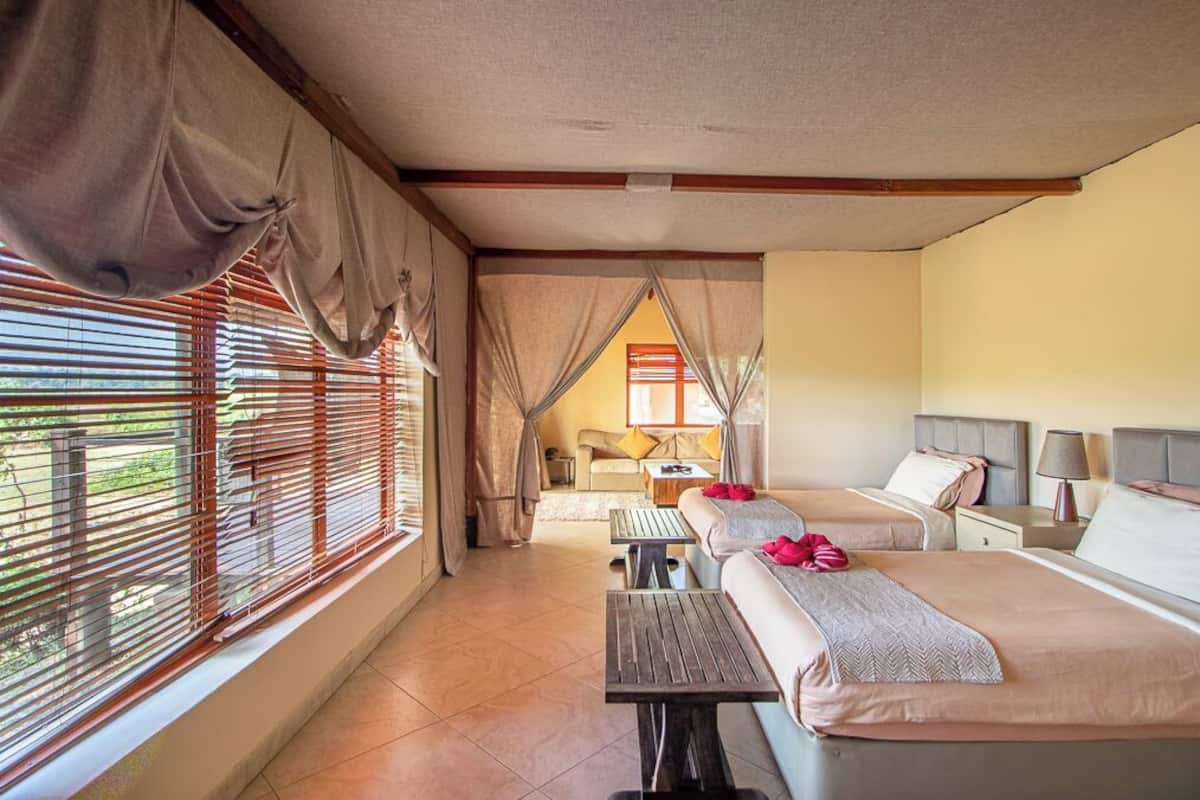A hotel room with two beds, a small table, and a view of the outdoors through large windows with blinds.