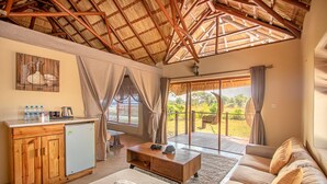 A spacious living area with a wooden ceiling, a sofa, a coffee table, a small kitchen with a refrigerator, and a large sliding glass door leading to an outdoor patio. - Mopani Safari Lodge (South Luangwa National Park)