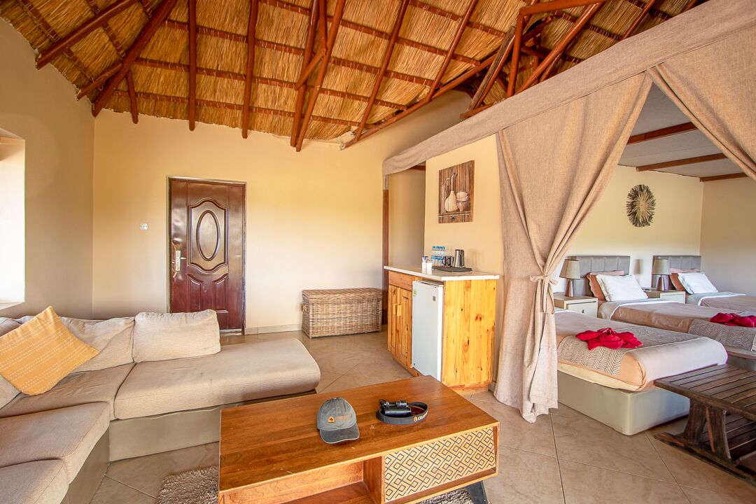 A room with a thatched roof, a sofa, a wooden table, a kitchenette, and a bed with red bedding.