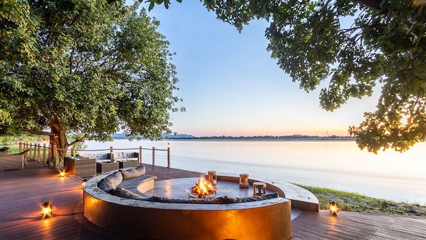 A circular outdoor seating area with a fire pit, surrounded by wooden deck and lanterns, overlooking a calm body of water. - Mopani Safari Lodge (South Luangwa National Park)