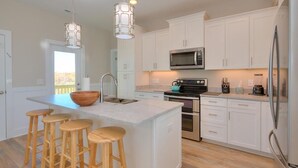 A modern kitchen with white cabinets, a central island with stools, stainless steel appliances, and pendant lighting. - Expansive 5BR Oceanview Home in Oak Island with Pool, Sleeps 16 & Dog Friendly! (Oak Island)