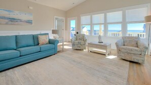 A spacious living room with a blue sofa, two patterned armchairs, a small table, and a large window with blinds. - Expansive 5BR Oceanview Home in Oak Island with Pool, Sleeps 16 & Dog Friendly! (Oak Island)