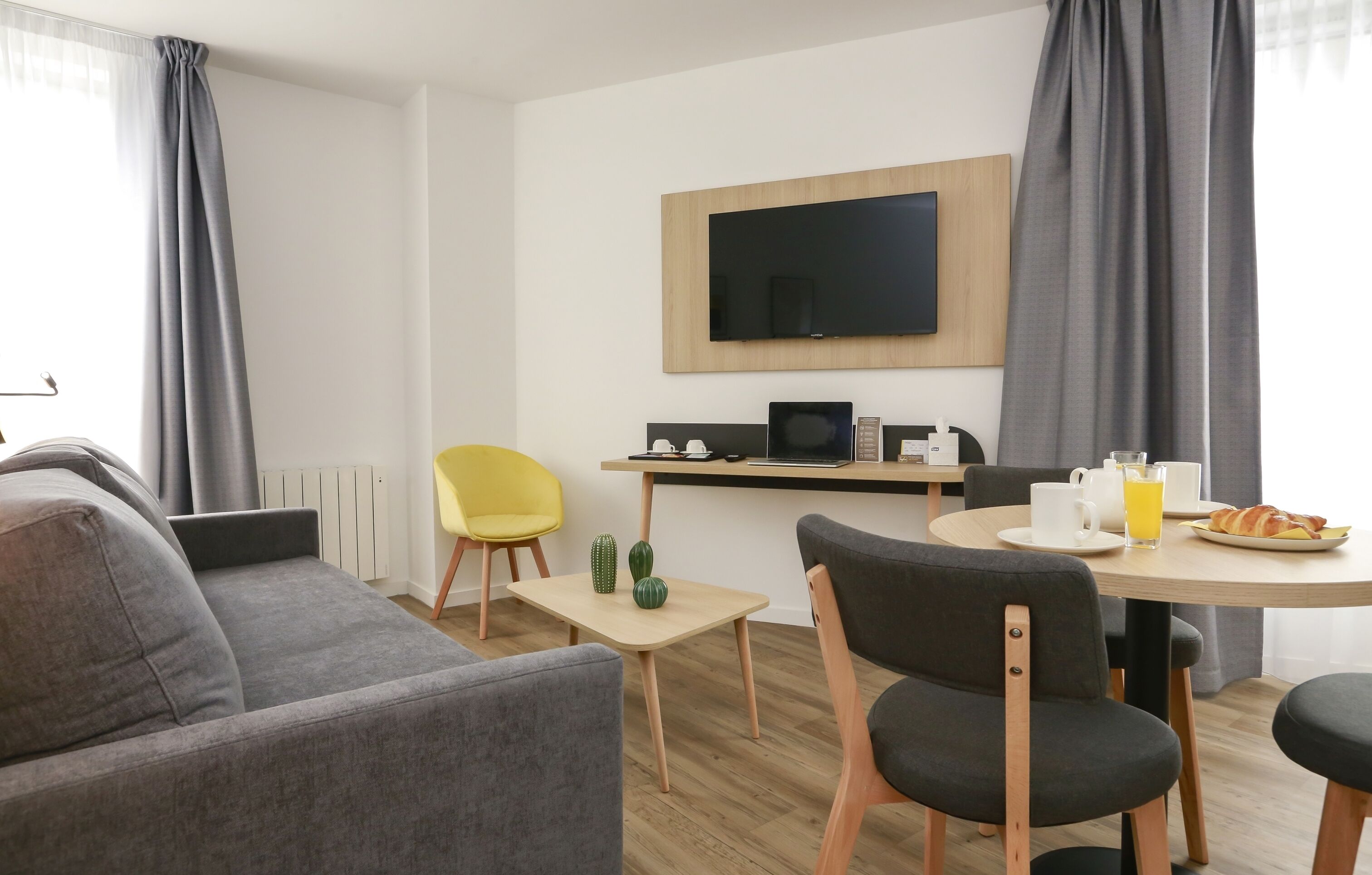 Ett modernt hotellrum med en soffa, ett matbord med stolar, en platt-TV som är monterad p å väggen och ett litet sidobord med en kaffebryggare och ett croissant.