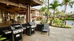 An outdoor patio with two portable grills, seating, and a pool area surrounded by tropical plants and palm trees. - Private townhome near Hawaii's best beaches (Waikoloa)