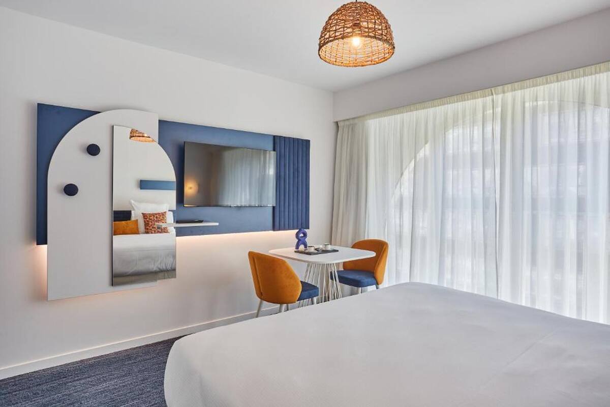 A hotel room with a bed, a dining table with chairs, a TV, and a large window with curtains.