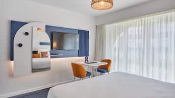 A hotel room with a bed, a dining table with chairs, a TV, and a large window with curtains.