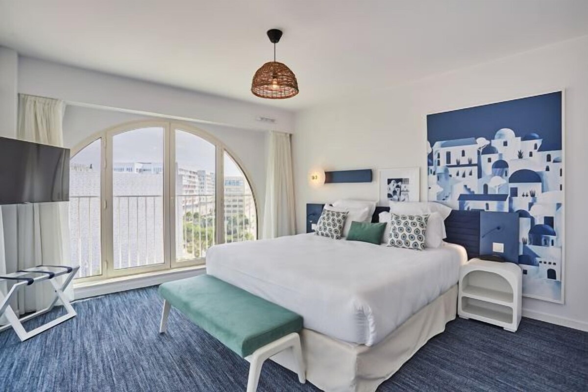 Habitación de hotel con una cama grande, un banco, un televisor y vistas a edificios a través de un ventanal.