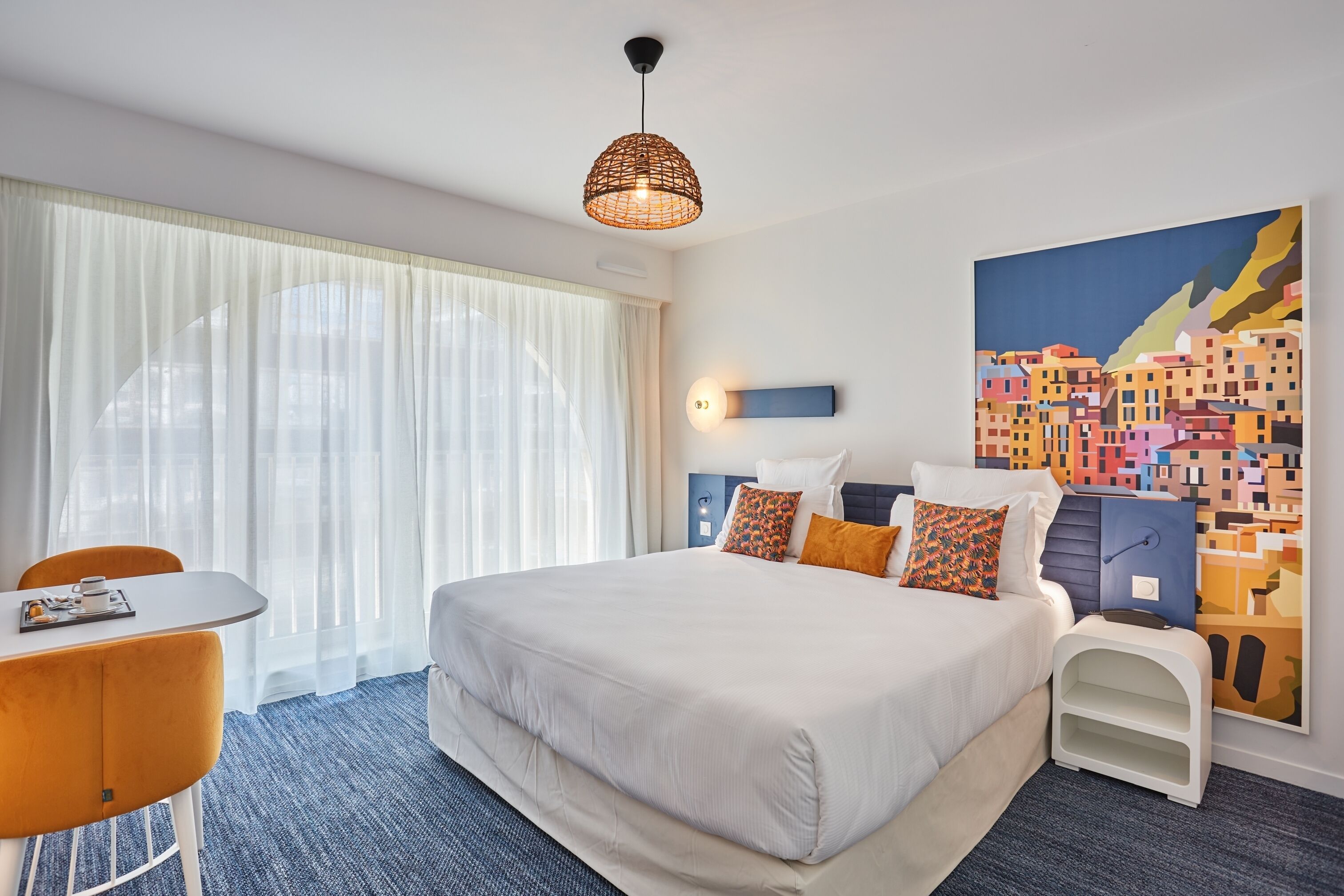 Habitación de hotel con una cama grande, una mesa de comedor, una silla, una mesita de noche, una lámpara y un cuadro en la pared.
