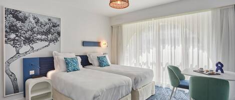 A hotel room with two beds, a desk, a chair, a hanging light fixture, and a large window with sheer curtains.