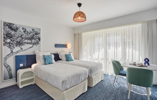 A hotel room with two beds, a desk, a chair, a hanging light fixture, and a large window with sheer curtains.