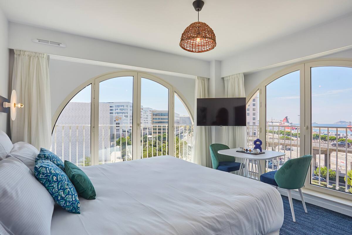 A hotel room with a large bed, a TV, a small table, and chairs. There are large windows with a view of the city.