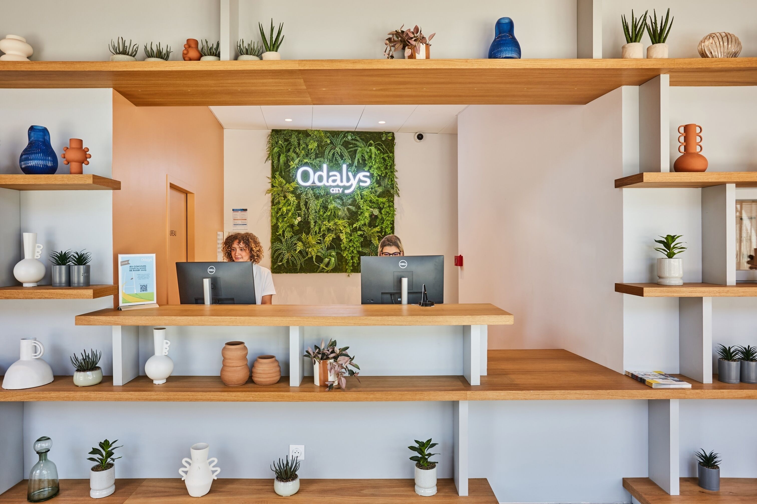Balcão de recepção com dois computadores, rodeado por prateleiras com plantas e objetos decorativos.