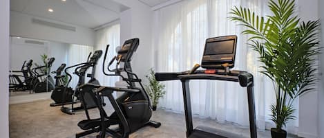 A gym with treadmills, stationary bikes, and an elliptical machine. There are large windows with sheer curtains and a potted plant.