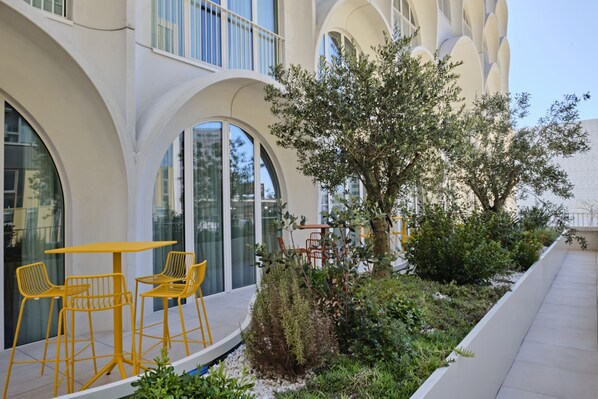 A modern building with large arched windows, outdoor seating with yellow chairs, and a well-maintained garden area.