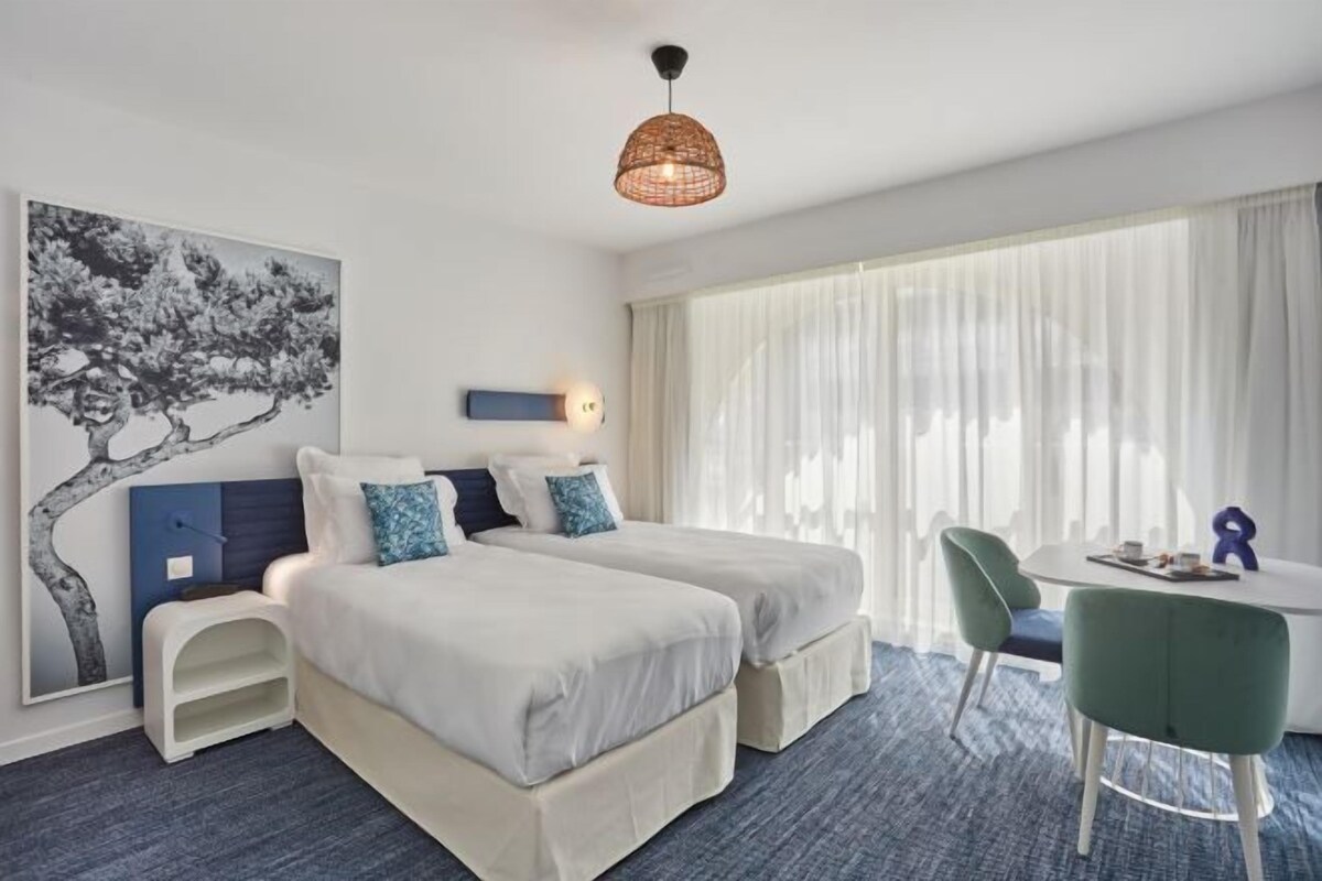 A hotel room with two beds, a desk, and a chair. There is a large window with sheer curtains and a hanging light fixture.