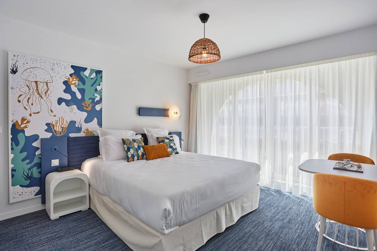 A modern hotel room with a large bed, a side table, a small desk, a chair, and a large window with sheer curtains.