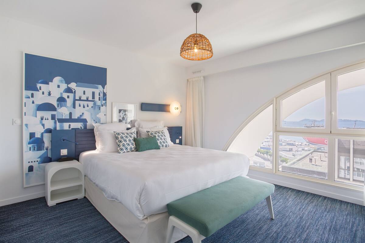 A modern hotel room with a large bed, a view of the city through large windows, a wall art of a blue and white cityscape, and a hanging light fixture.