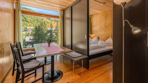 A modern room with a wooden ceiling, a bed with white bedding, a table with chairs, and a view of a forested area. - Holzboxen Planneralm (Irdning-Donnersbachtal)