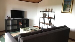 A living room with a flat-screen TV on a black stand, a glass coffee table, a dark sofa, a bookshelf with decorative items, and a framed painting on the wall. - ETNA VIEW-CATANIA-VALVERDE (Maugeri)