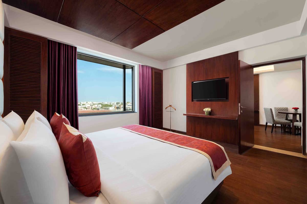 Ein Hotelzimmer mit einem großen Bett, einem an einer Holzvertäfelung montierten Fernseher, einem Essbereich mit Tisch und Stühlen sowie Blick auf die Stadt durch das Fenster.