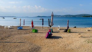 A pebbled beach with umbrellas, chairs, and luggage, overlooking calm waters and distant hills.