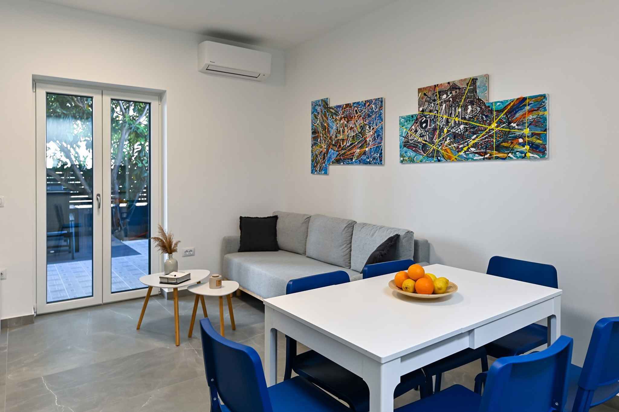 A modern living room with a white sofa, blue chairs, a white table with fruit, and abstract paintings on the wall.