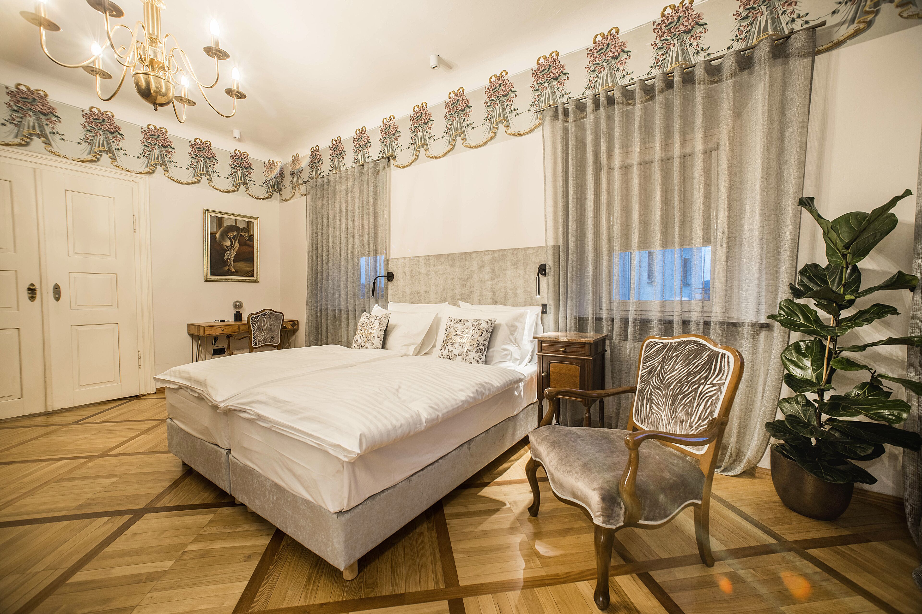 A hotel room with a large bed, a wooden chair, a nightstand, a potted plant, and a chandelier.