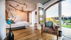 Una camera da letto con un letto, una sedia, un vaso con rami e un'ampia finestra vista sul verde.