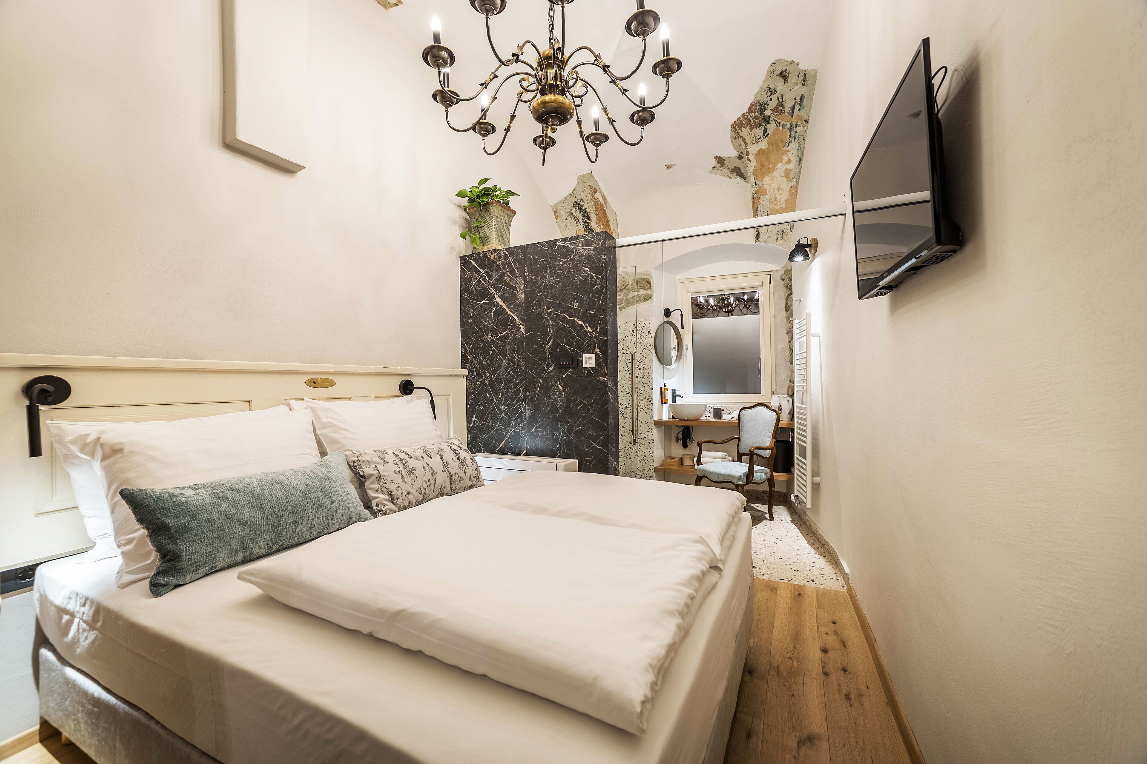 A bedroom with a bed, a chandelier, a TV mounted on the wall, and a bathroom visible through an archway.