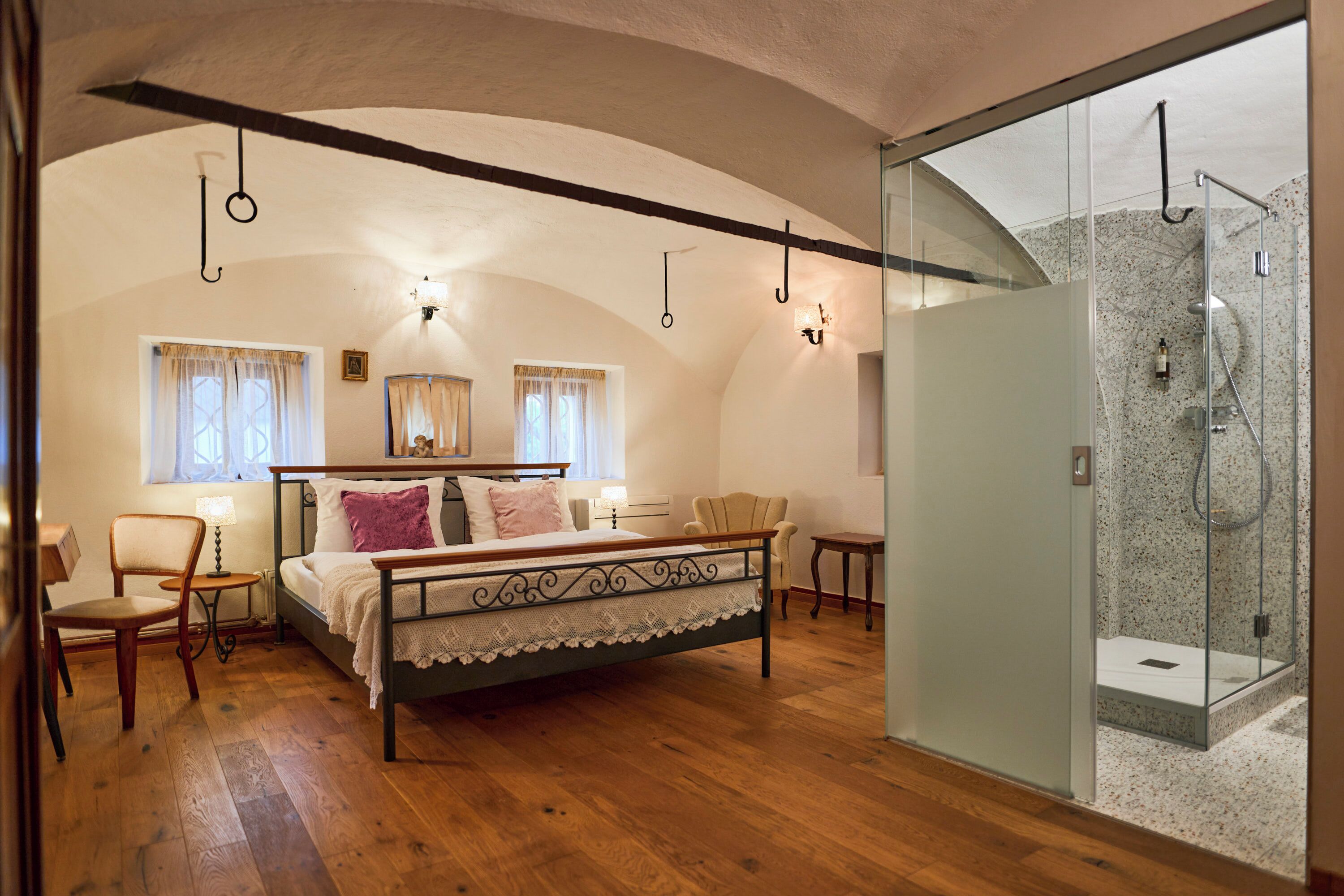 A bedroom with a bed, a chair, a small table, a nightstand, a glass shower enclosure, and a window with curtains.