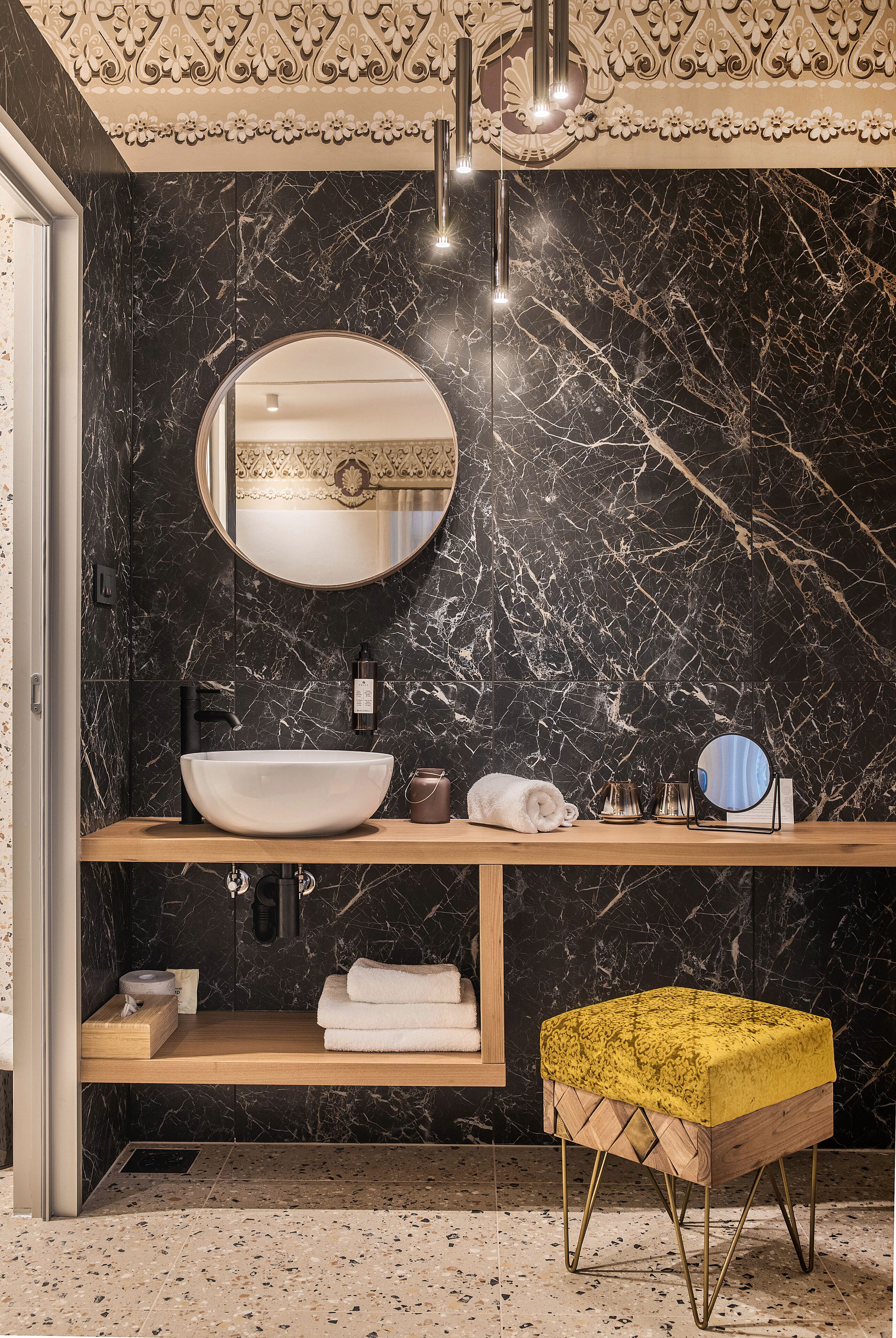 Une salle de bain moderne avec un mur en marbre noir, un miroir rond, un meuble-lavabo en bois et un pouf jaune.
