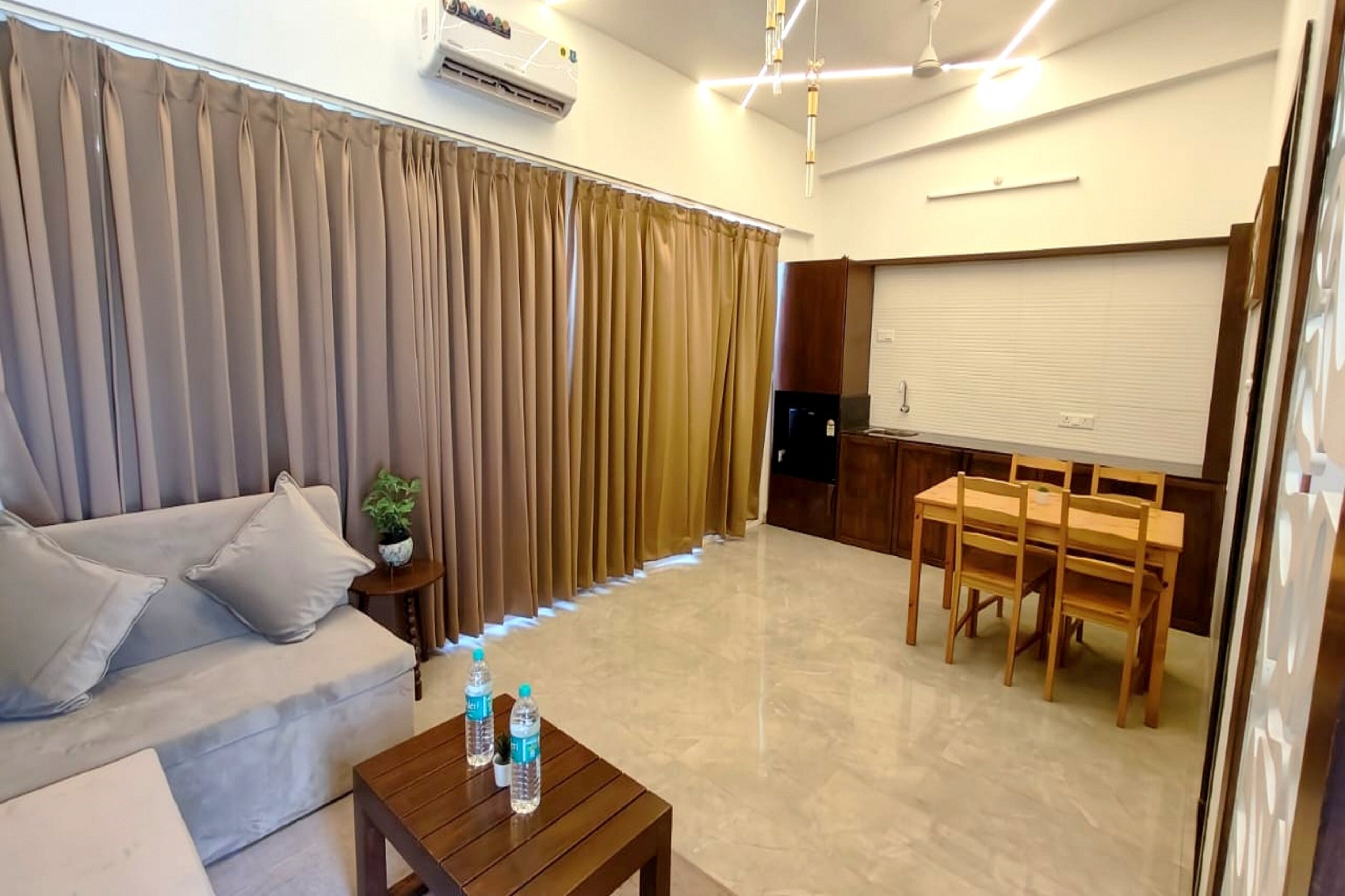 Uma sala de estar com um sofá, uma mesa de centro com garrafas de água, uma área de jantar com mesa e cadeiras, e uma janela fechada com persianas.