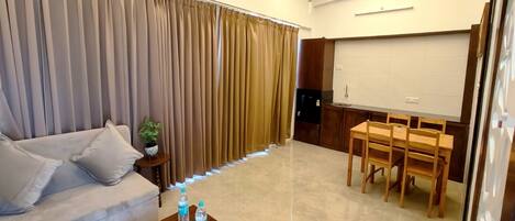 Uma sala de estar com um sofá, uma mesa de centro com garrafas de água, uma área de jantar com mesa e cadeiras, e uma janela fechada com persianas.