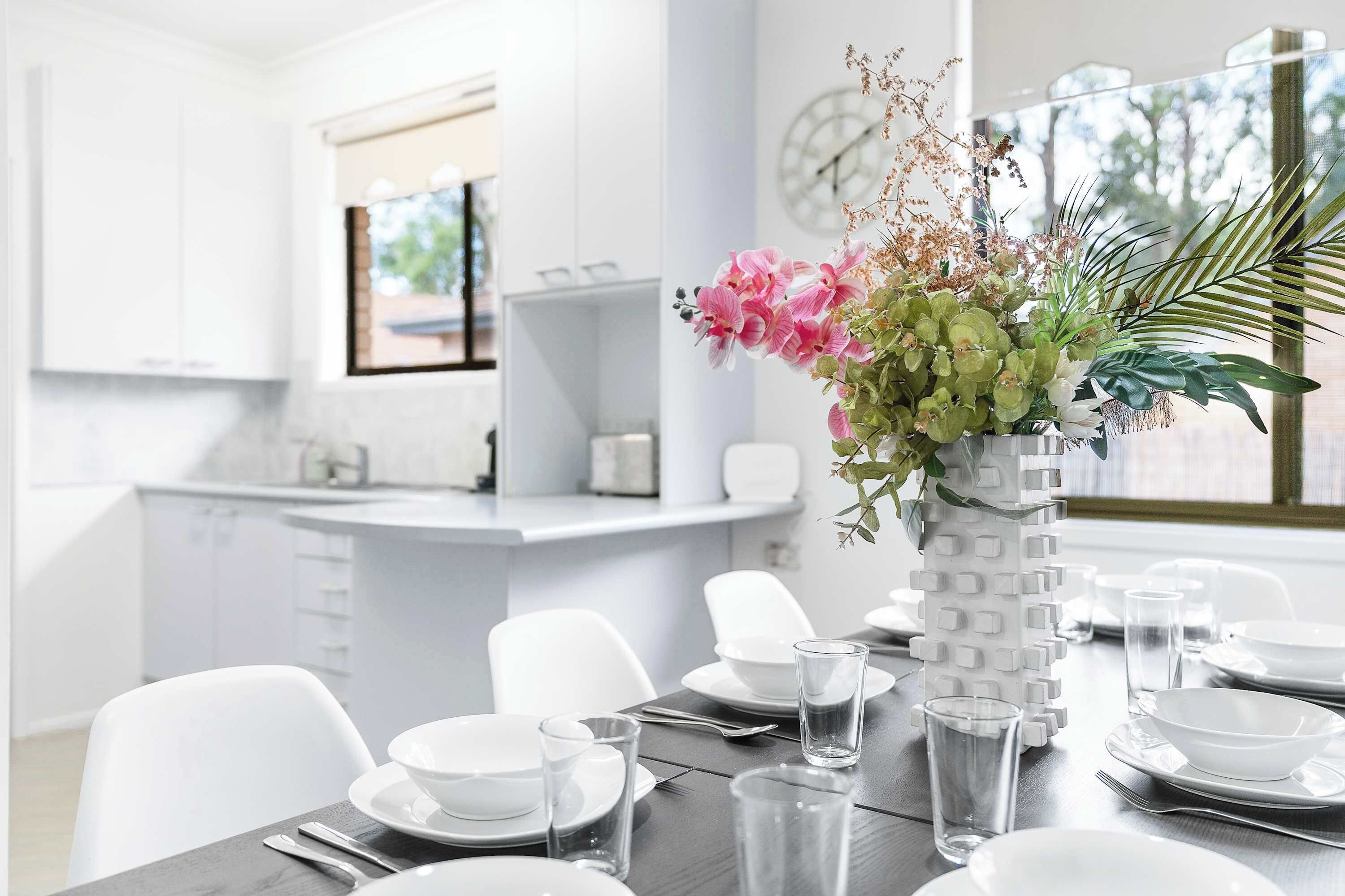 A modern kitchen with white cabinets, a dining table set with plates, glasses, and cutlery, and a decorative vase with flowers.