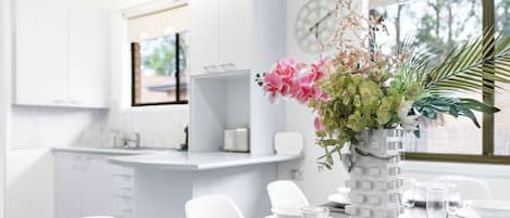 A modern kitchen with white cabinets, a dining table set with plates, glasses, and cutlery, and a decorative vase with flowers.