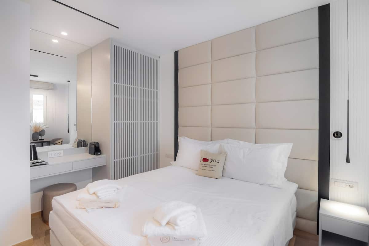 A modern hotel room with a large bed, a bedside table, a built-in wardrobe, a bathroom with a mirror, and a small seating area.