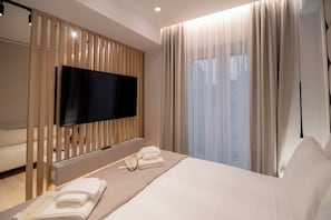 A modern hotel room with a large bed, a flat-screen TV mounted on a wooden panel, a bedside table with towels, and a large window with sheer curtains.