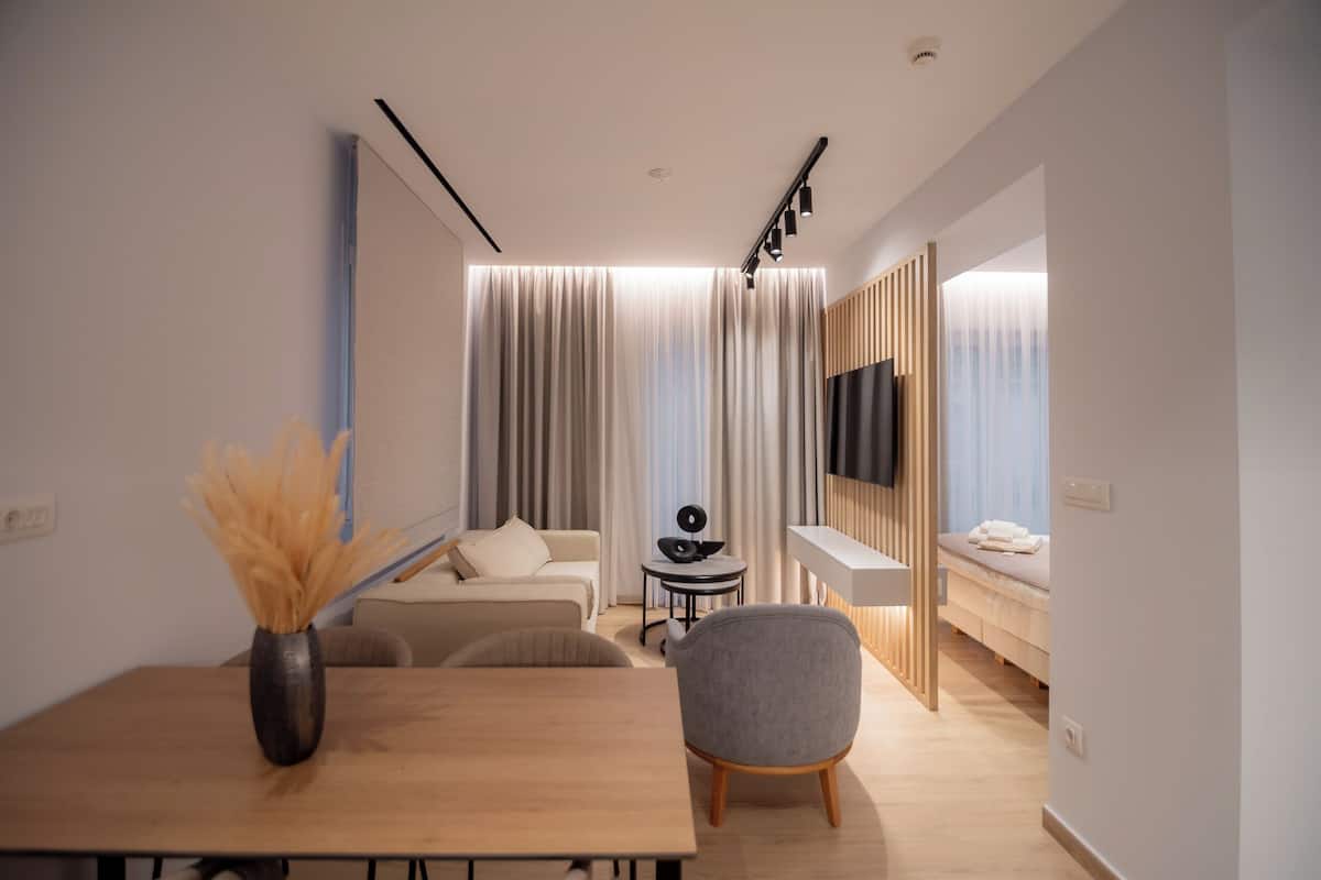 A modern hotel room with a wooden dining table, a grey armchair, a small round table with a decorative item, a sofa, a bed with a wooden headboard, and a mounted TV.