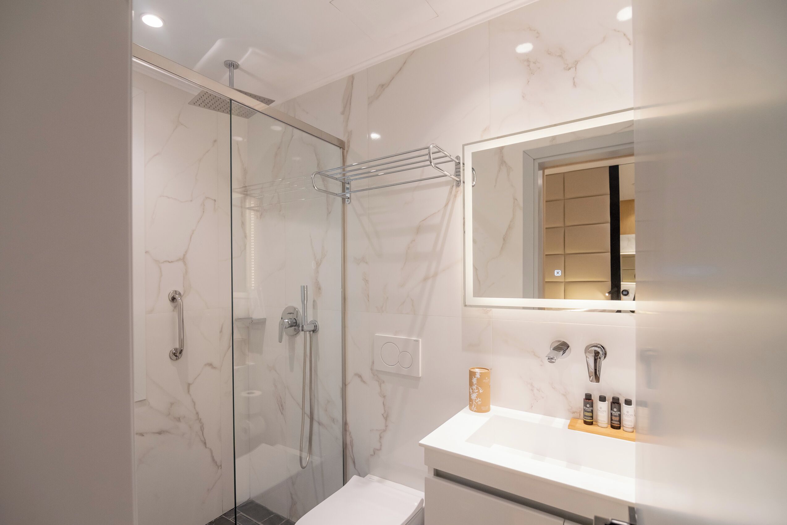 A modern bathroom with a glass-enclosed shower, a white sink with a marble countertop, and a mirror with a wooden shelf holding toiletries.