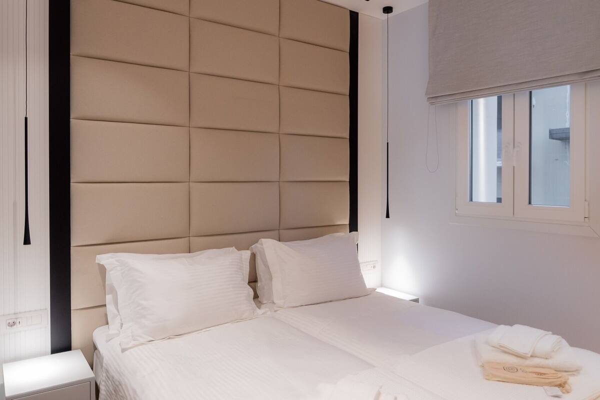 A modern bedroom with a large bed, a bedside table, and a window with blinds.